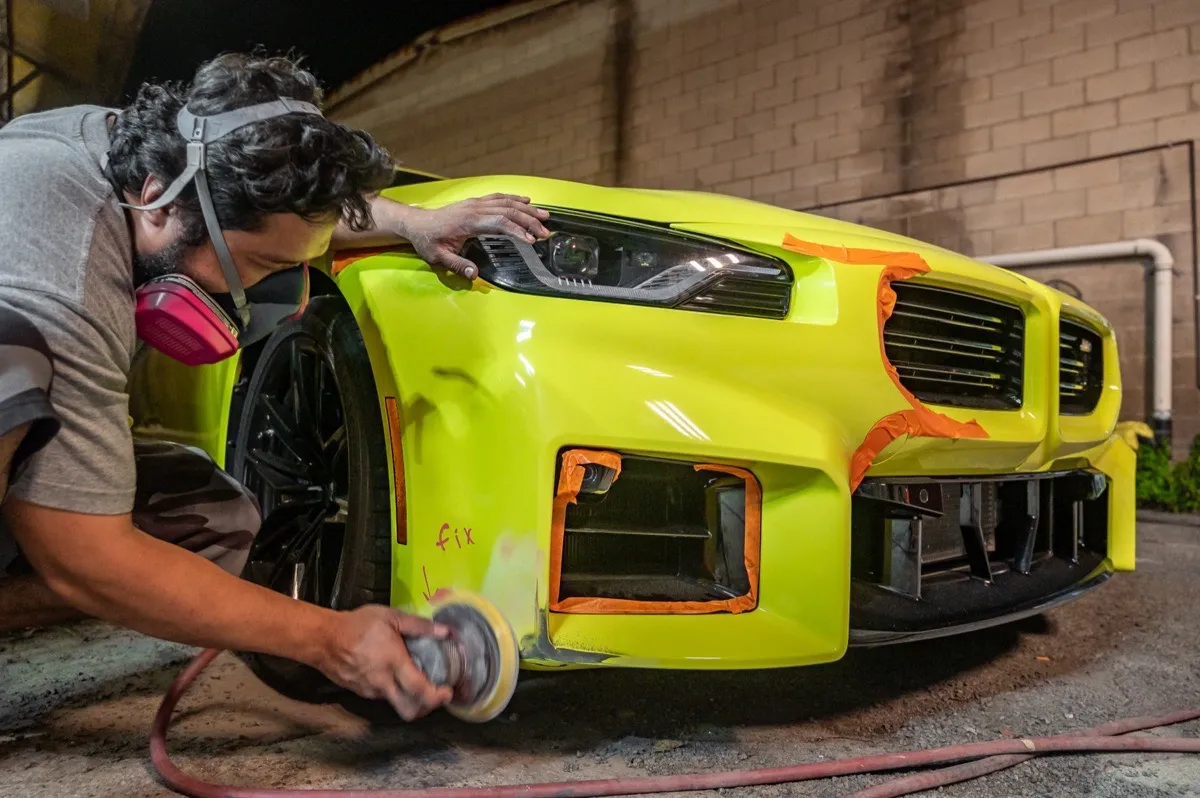 Technician sanding the marked bumper corner of a yellow BMW M2